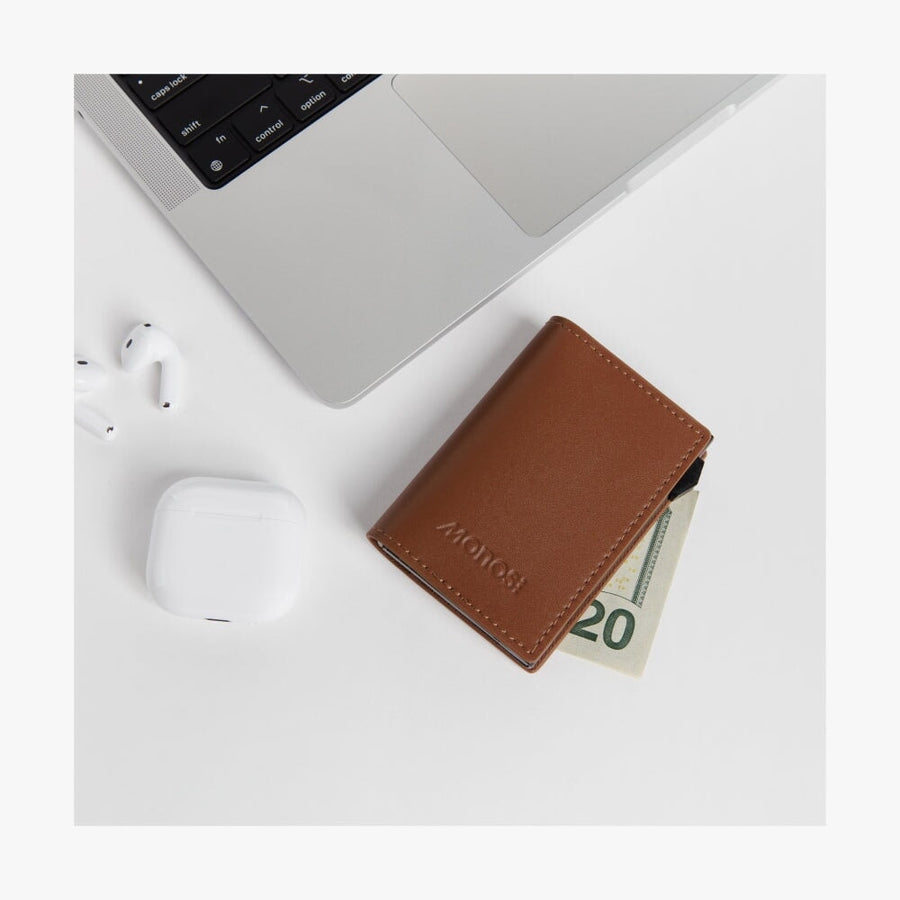 Mahogany / Black | Slim Wallet Pro in Mahogany / Black placed on a white desk beside a laptop, wireless earbuds, and their charging case. A folded US twenty-dollar bill is partially visible in the wallet.